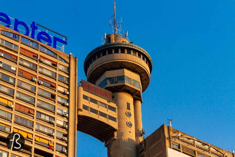 The building got the nickname Genex Tower since one of the towers was occupied but the Genex Group for years. Now, it's abandoned, but their Zepter logo can still be seen over the commercial side of the building. The smaller tower is a residential building with some shops on the ground floor and a few people passing by.